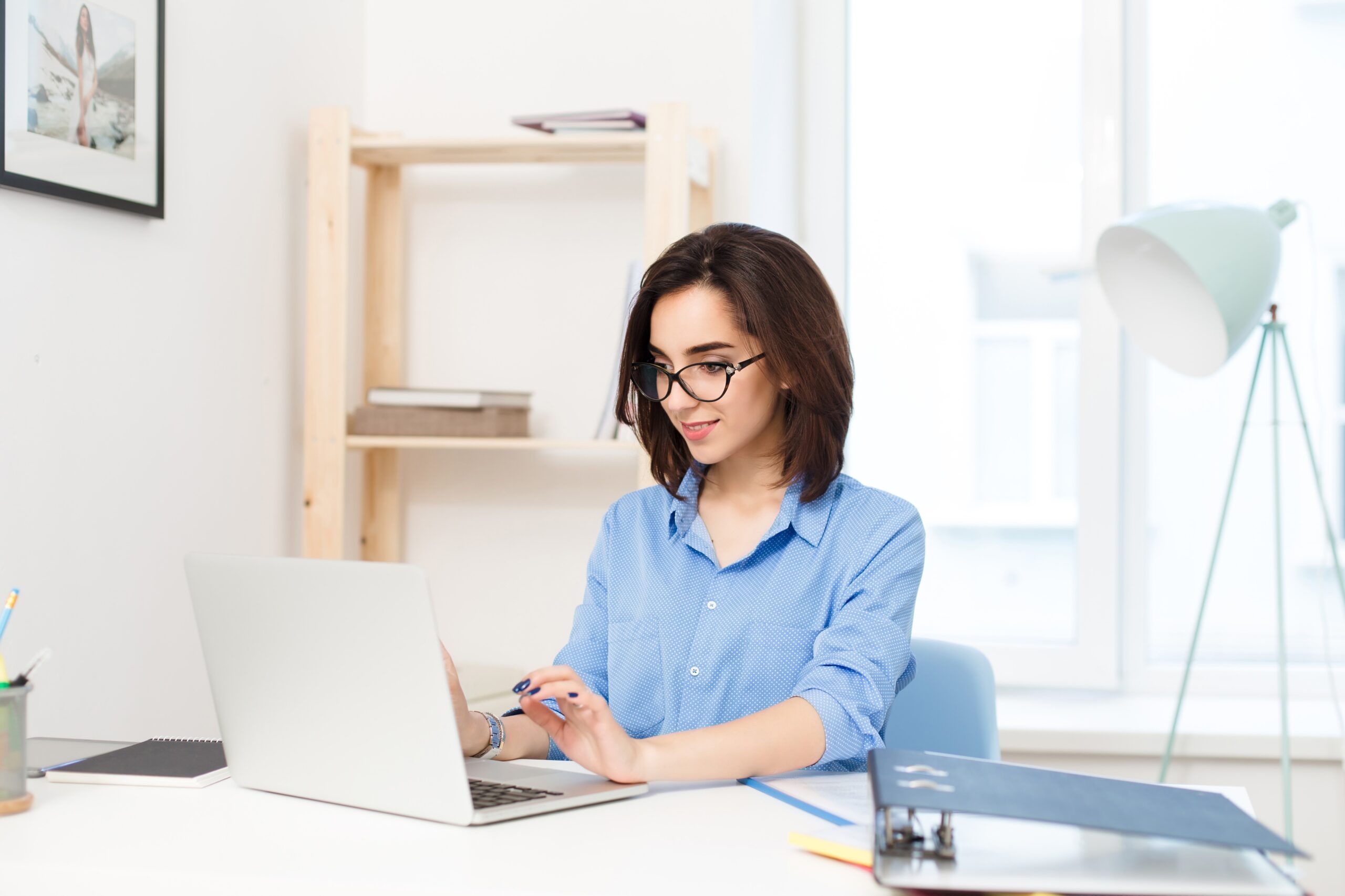 A pretty brunette girl in a blue shirt sitting at the table in office. She is typing on laptop and looks happy